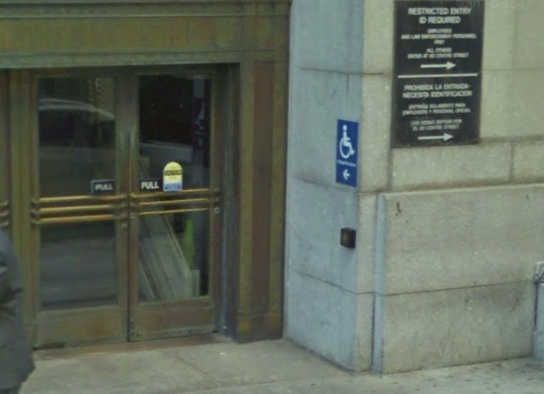 A double door entrance with ADA signage posted on the building to the right. There is a push button located on the right side of the building under the ADA signage. The entrance is accessed on the street level.
