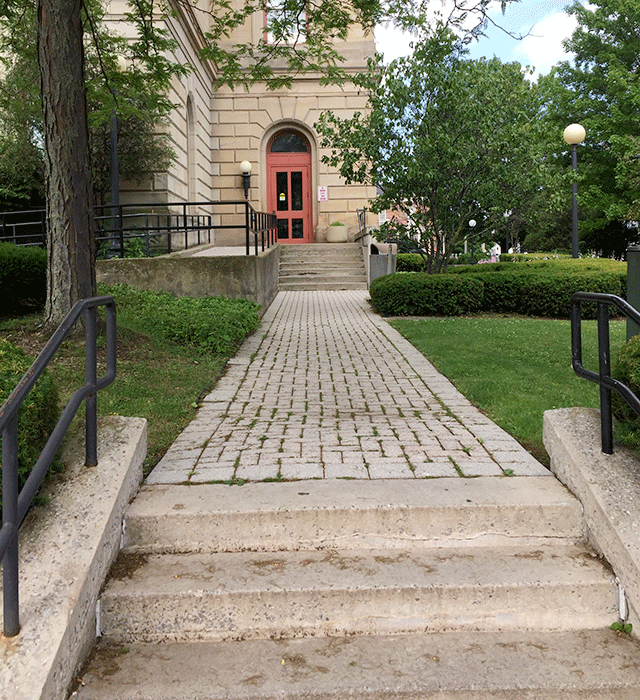 The entrance from the view of a set of stairs that lead to a walkway and another set of stairs to the entrance. The walkway is between a grassy area with trees. To the left is a ramp that also leads to the same entrance.