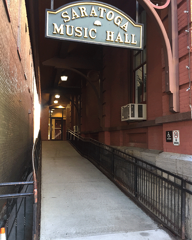 A ramp leading to a single door. There is ADA signage and a no smoking sign on the right hand side of the building. Above the ramp is a sign that says 'Saratoga Music Hall.'