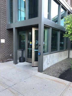 An open door to the courthouse. The entrance is accessed from the sidewalk and there is a push button to open the door to the right of the door located on a column of the building.