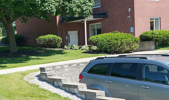 A walkway that leads to the ADA-accessbile entrance. There is signage on the side of the facility and the entrance is a single door to the left of the walkway. There is a lawn with trees and bushes aurrounding the walkway area.