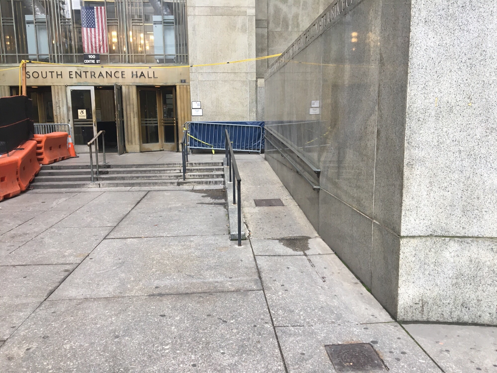 A walkway leading to steps. The steps lead to two revolving doors separated by double doors entry. The double door entry is open. To the right of the walkway is a ramp that leads to the entrance area.