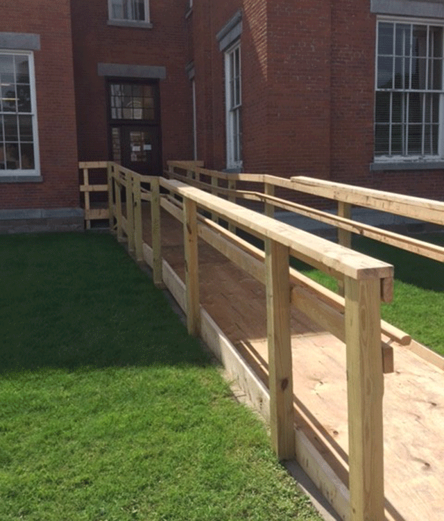 A wooden ramp crossing a patch of grass. The ramp leads to a single door entrance.