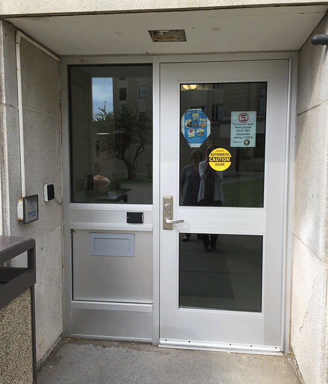 A single door entrance to the courthouse. There is a caution sign on the door warned that the door is automatic. The push button to open the door is on the left side up against the wall. There is also a bell on the same wall as the button.