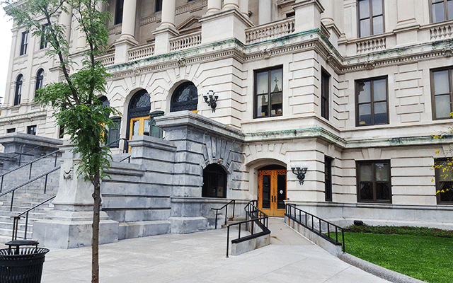 A view of the courthouse from the street level. There is a large set of stairs that leads to multiple sets of doors. To the right on the sidewalk is a ramp that leads to a single set of doors. The single set of doors are also accessed by three steps to the left of the ramp.