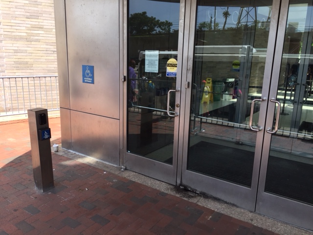A  single door with a set of double doors to the right. The entrance is accessed on street level. There is a pole with a push button on the sidewalk toward the left hand side.