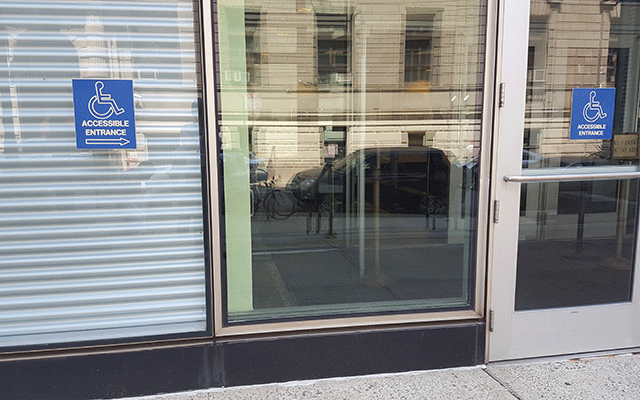 A window with ADA signage pointing to a single door. The door also has ADA signage and is located on the street level.