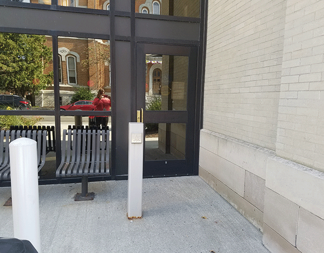  Courthouse with a single door to the far right. To the left is a push button that will open the door. There is outdoor seating to the left of the door and a push button.
