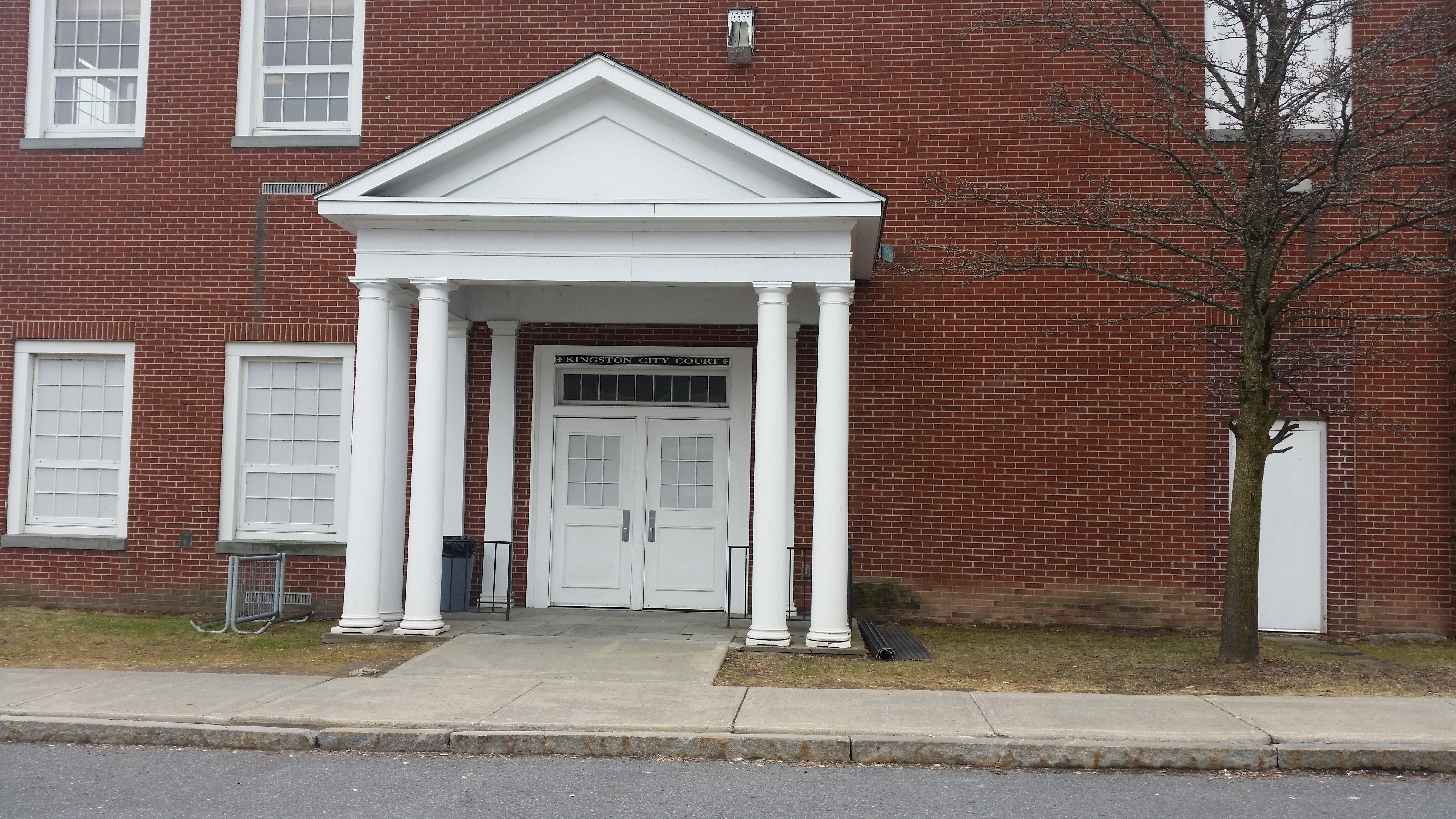 This is the entrance to the Kingston City Court. There are two doors that are entered through directly from the sidewalk. The entryway is covered by a peaked area held up by large columns.