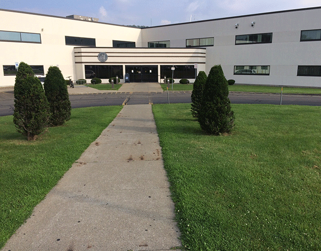 A walkway through a grassy area with bushes leading to a crosswalk. Beyond the crosswalk is the entrance to the facility that can be accessed on the street level.