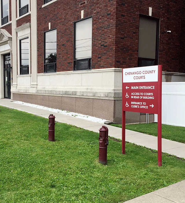 The Chenango County Courthouse main entrance to the facility. There is a ramped walkway that leads directly to the doors. There is also a sign in the grassy area that points in the direction to other offices within the facility.