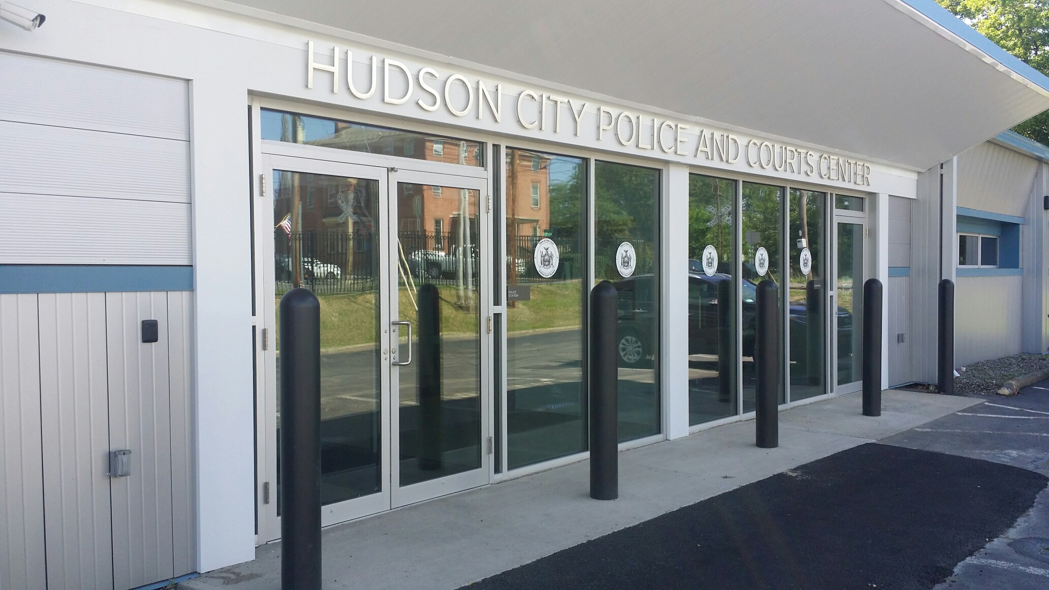 Entrance to the Hudson City Police and Courts Center. There's a set of doors on street level to the left of the entrance. There are also black poles coming up from the ground that are 6 to 10 feet apart. These poles are lining the length of the entry area.