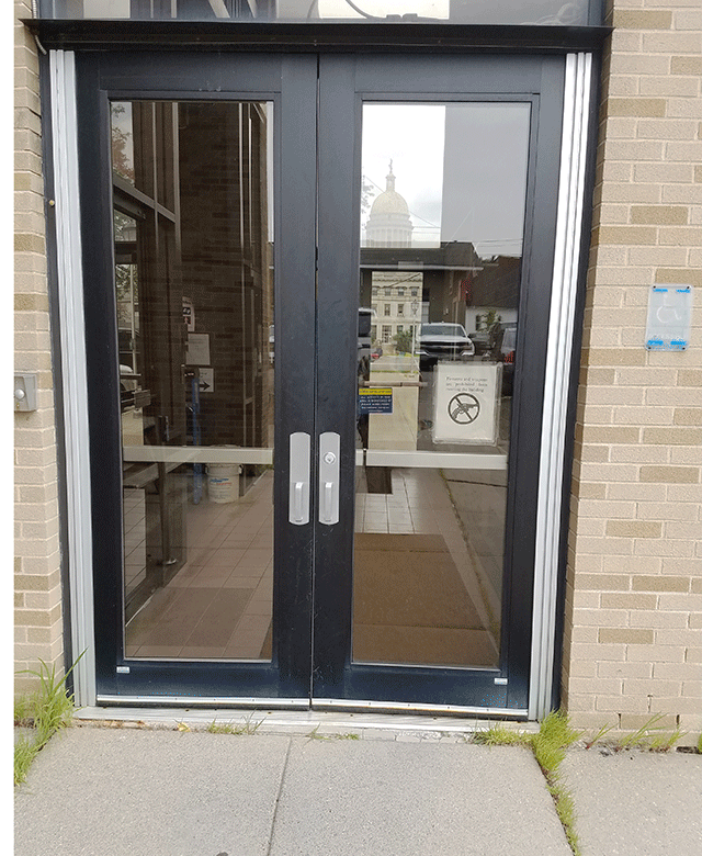 The photo is of double doors that can be accessed from the sidewalk. There is an ADA-accessible sign on the right side of the doors and a buzzer on the left side  of the doors. There is also a  no weapon sign posted directly on the right door.