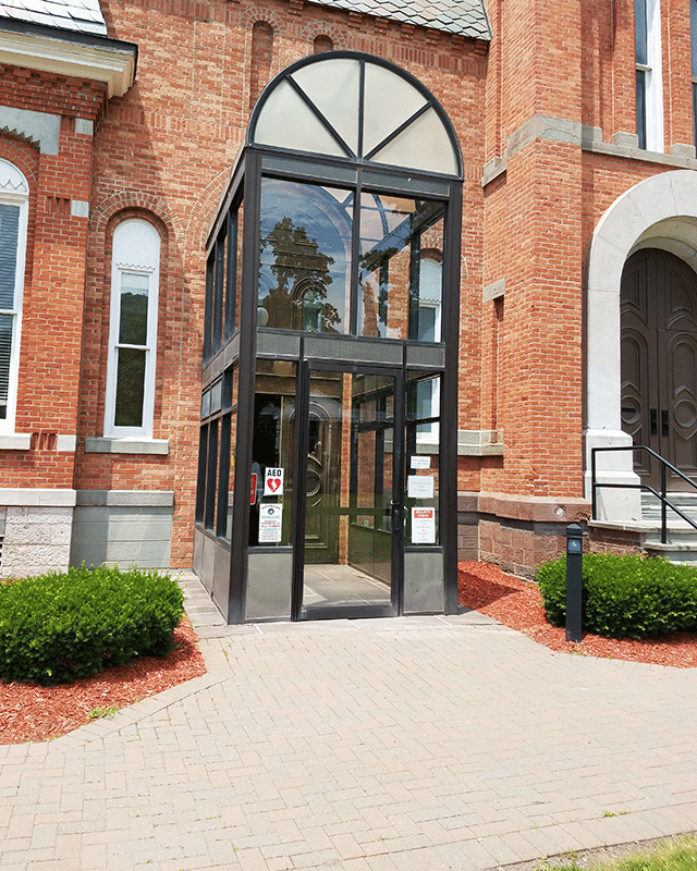 A walkway that leads to the accessible entrance. There is a push button on a black post to open the door of the walkway located on the right side.
