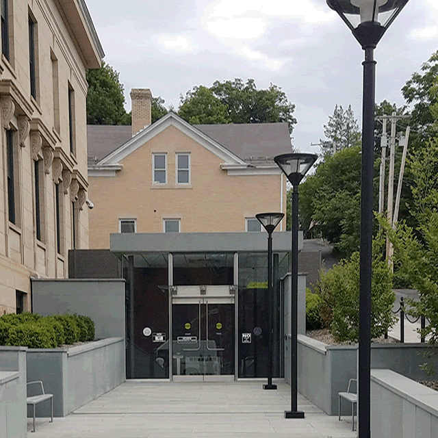 A long walkway with a bench on the left and light posts to the right. Straight ahead is a set of double doors accessed directly from the walkway.