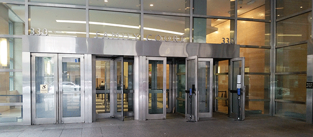 The multiple door entrance to Family Court from street level. Double doors marked with ADA signage to the far left, followed by a revolving door and then two additional double doors. Each of the double doors on the right have one door open.