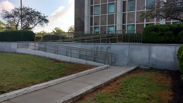 A walkway between two grassy areas. The walkway leads to a ramp that leads to the court facility behind it.
