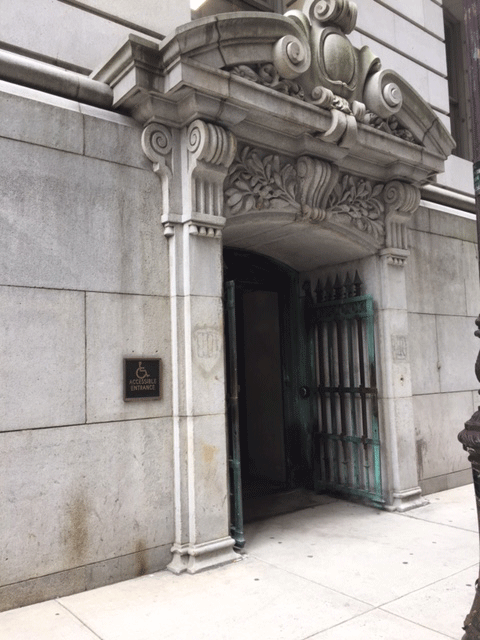The entrance to the courthouse that has two opened metal gates. The entrance is accessd from the street level. There is ADA signage posted against the building to the left of the entrance. There is a push button to the right of the entrance doors.