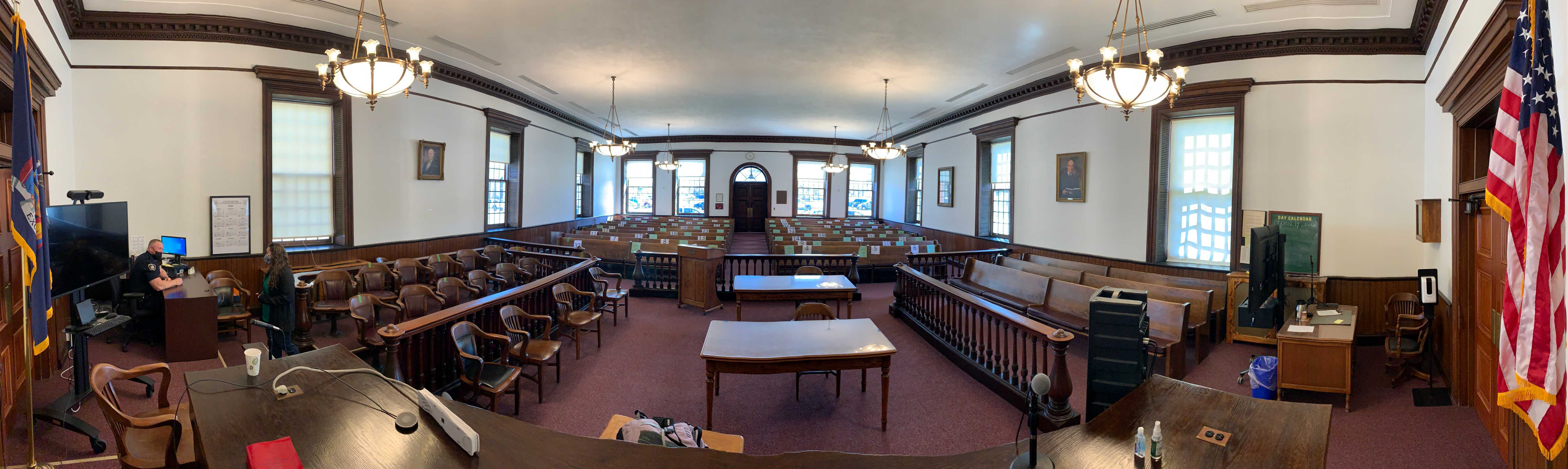 An old-fashioned courtroom with wooden furniture has been modernized with two large TV screens and CPUs on rollable stands (one computer is connected to a videoconferencing camera), a speaker, and microphones. The panorama is taken from the viewpoint of the judge's bench, facing the gallery.