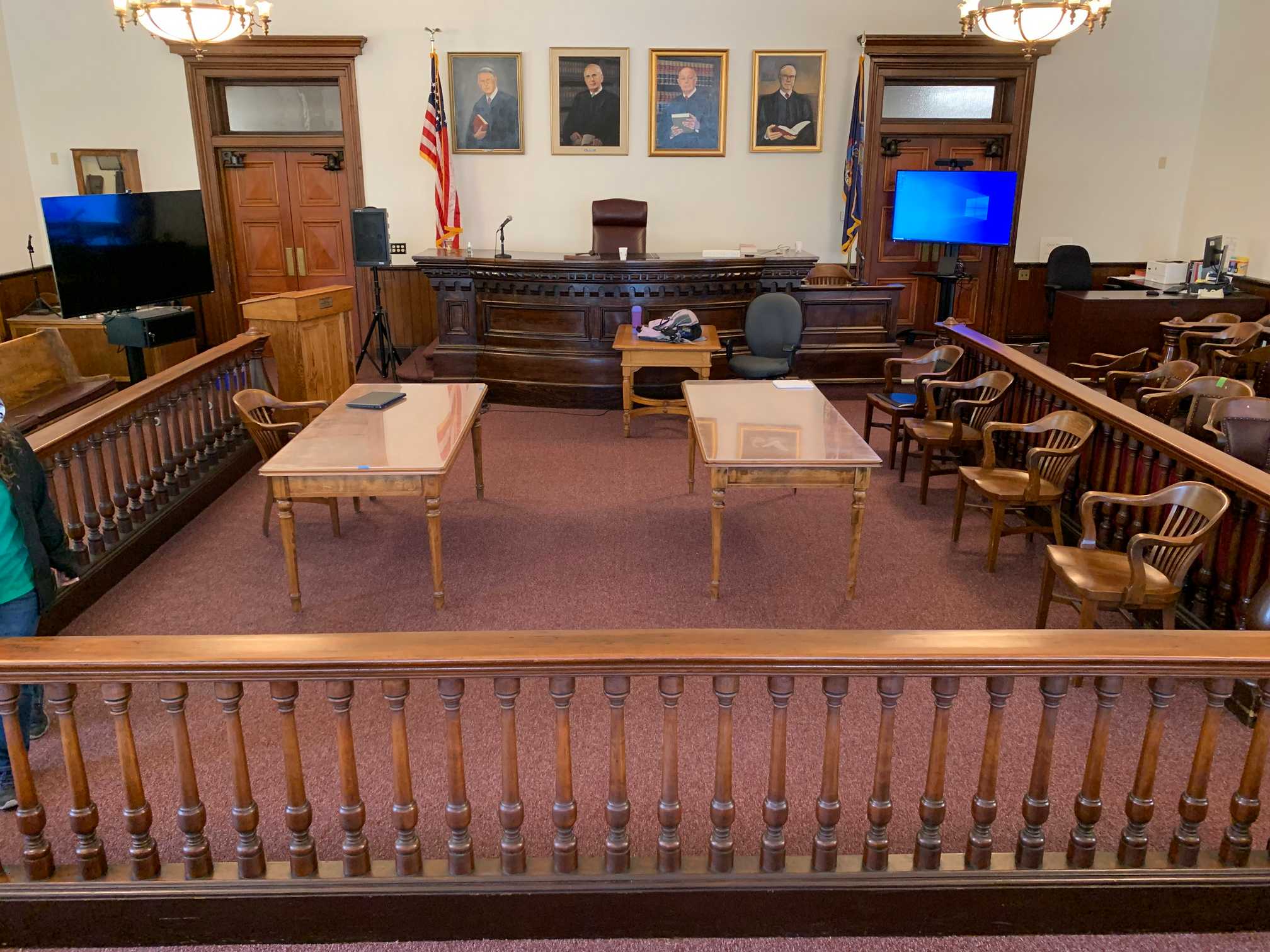 An old-fashioned courtroom with wooden furniture has been modernized with two large TV screens and CPUs on rollable stands (one computer is connected to a videoconferencing camera), a speaker, and microphones. The picture is taken from the viewpoint of the gallery, facing the judge's bench.