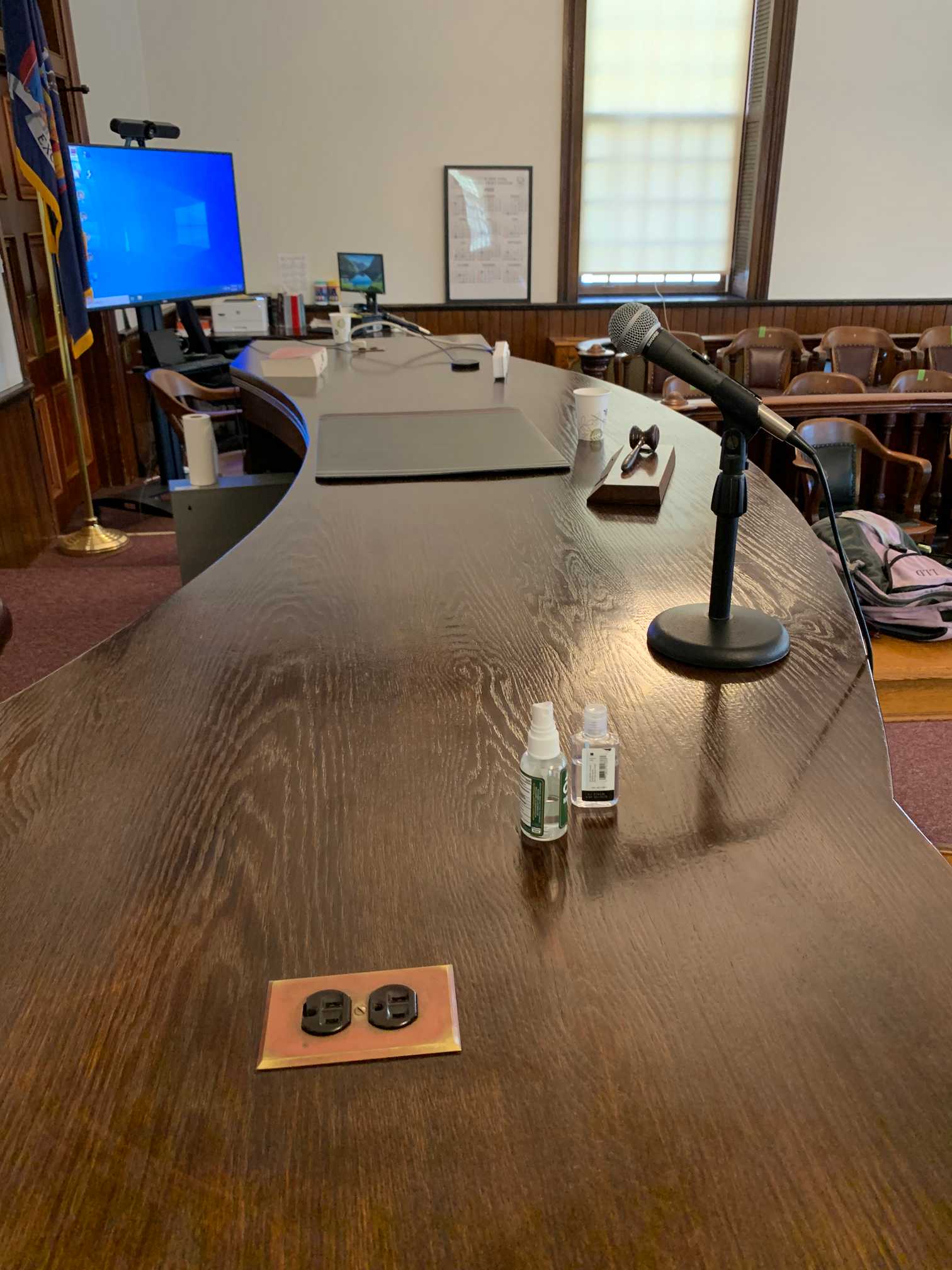 An old-fashioned courtroom with wooden furniture has been modernized with various forms of technology, but only a videoconferencing computer on a movable stand, microphones, and an outlet built into the judge's bench are visible. The picture is taken from the right side of the judge's bench, facing the left side of the bench (i.e. right and left from the perspective of the judge's bench, facing the gallery).